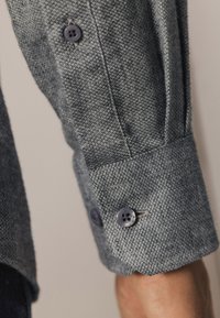 Grijze gestructureerde shirtstof met contrasterende paarse knopen en een detail aan de mouw, die een geweven patroon en strakke lijnen tentoonstelt.