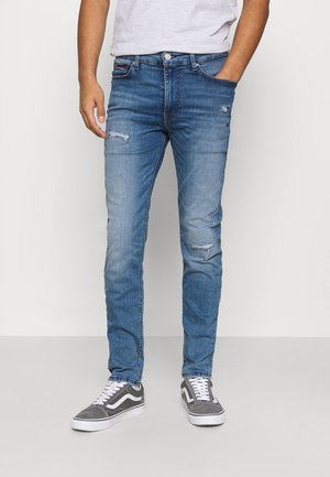 Mann trägt Slim-Fit hellblaue, abgenutzte Jeans und graue Schnürsneaker, steht mit einer Hand in der Tasche.