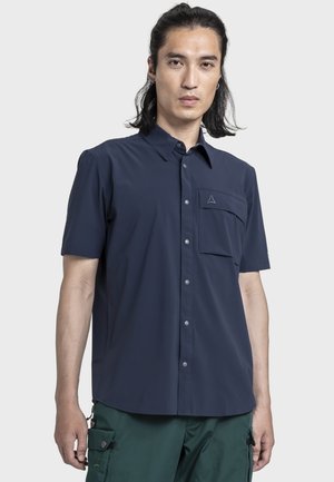 Mann mit schulterlangen Haaren, der ein marineblaues kurzärmliges Hemd mit Knöpfen und grüne Cargohosen trägt, steht vor einem schlichten Hintergrund.