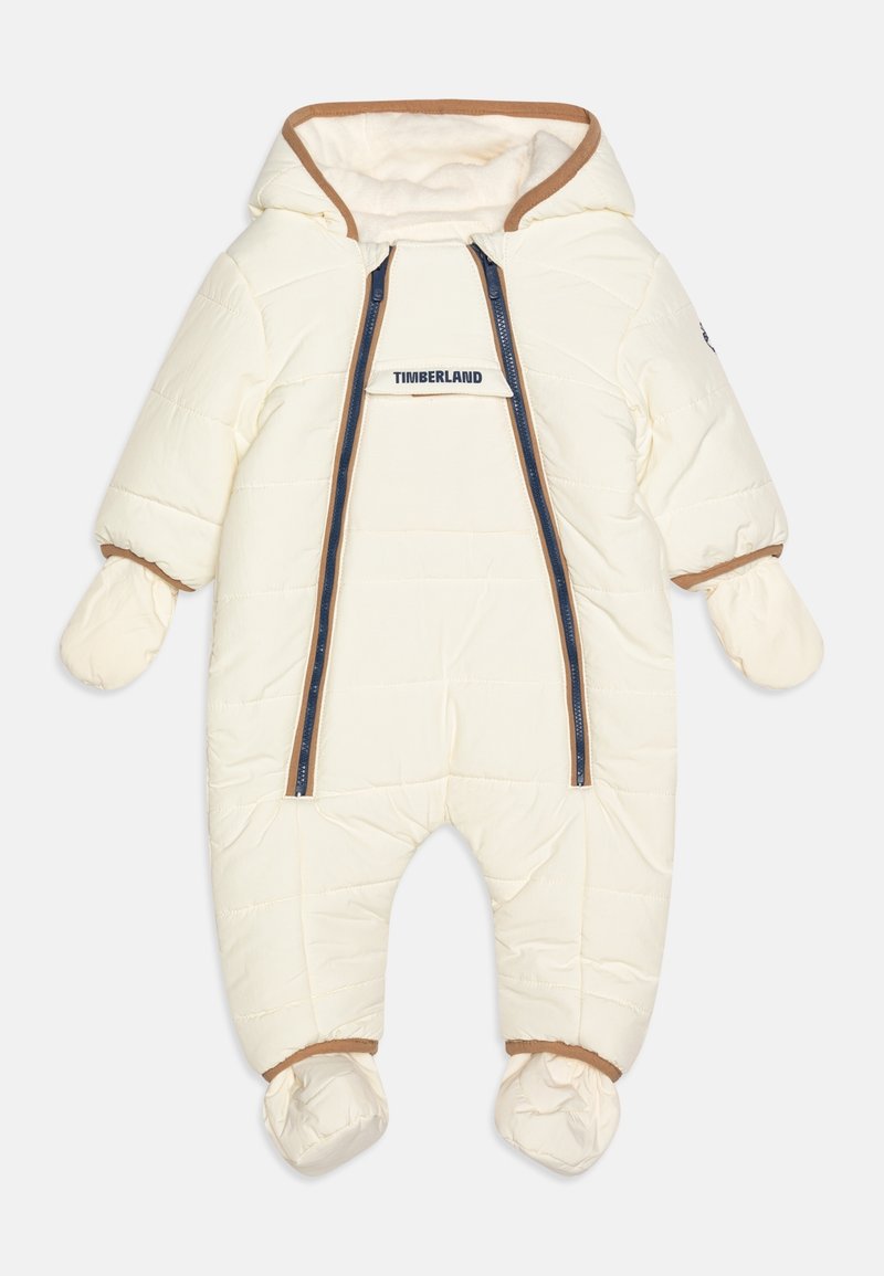 Combinaison de neige rembourrée pour bébé couleur crème avec moufles et chaussons attachés, bordure marron, double fermeture éclair devant et logo Timberland sur le rabat de poitrine.