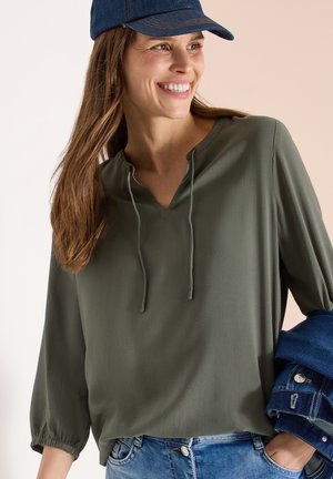 Smilende kvinde med langt brunt hår iført en blå kasket, olivengrøn bluse med snore og holder en denimjakke over armen.