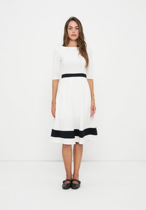 Femme debout portant une robe blanche longueur genou avec des bandes noires horizontales à la taille et à l'ourlet, associée à des chaussures noires.