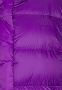 Violet puffer takki, jossa on kiiltävä, tikkikuvio. Etupuolella on logotunnuksia ja kevyt, eristetty muotoilu.