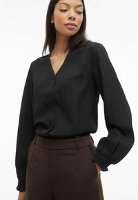 Melnā, garroku blūze ar V veida izgriezumu, vertikāliem svītrām un pogu aizdari. Piedurknes ar aproci un krokojuma detaļu.