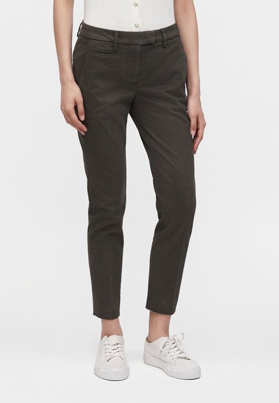 Personne portant un pantalon slim foncé coupé au-dessus de la cheville et des baskets blanches décontractées à lacets, debout devant un fond uni.