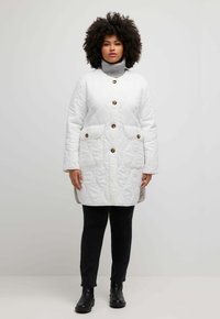 Bílý prošívaný kabát s hnědými knoflíky, dvěma předními kapsami, vysokým límcem a texturovaným povrchem; nošený přes černé kalhoty a kotníkové boty.
