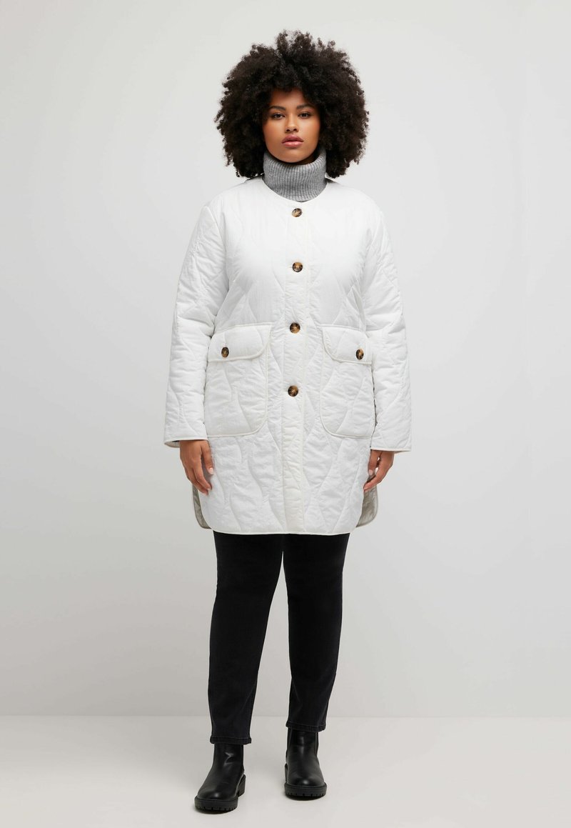 Bílý prošívaný kabát s hnědými knofl íky, dvěma předními kapsami, vysokým límcem a texturovaným povrchem; nošený přes černé kalhoty a kotníkové boty.