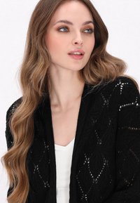 Cárdigan de punto negro con frente abierto, patrón de hojas intrincado y textura suave, usado sobre una blusa blanca.