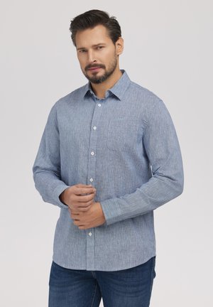 Mężczyzna z krótkimi ciemnymi włosami i brodą, ubrany w jasnoniebieską koszulę na guziki i ciemne dżinsy, trzymający ręce blisko talii.