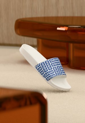 Balta šlepetė su mėlyna diržele, ant kurios kartojasi „HUGO“ logotipai, padėta ant šviesios kiliminės dangos šalia rudo permatomo kavos staliuko.