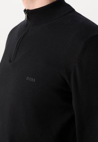 Gros plan d'une personne portant un pull en maille noir avec une courte fermeture éclair et un logo "BOSS" discret sur la poitrine.
