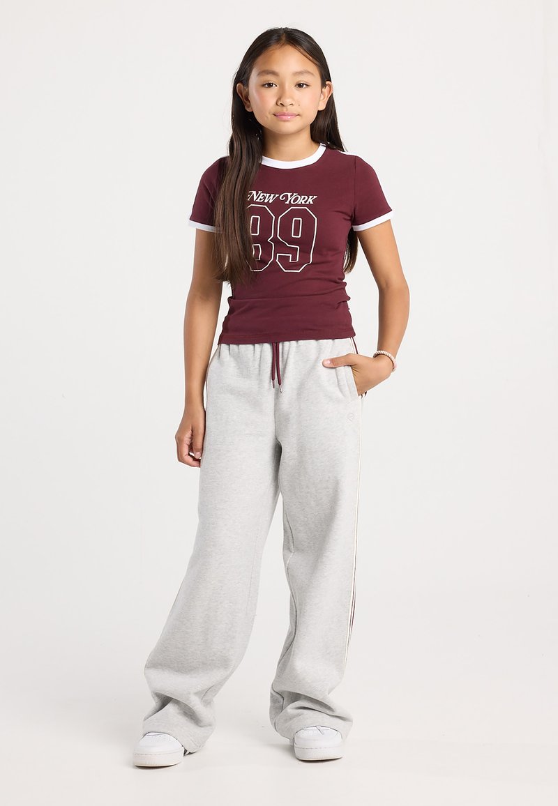 Fille aux longs cheveux noirs, portant un t-shirt marron "New York 89", un pantalon de survêtement gris clair, des baskets blanches et un bracelet au poignet droit.