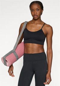 Μαύρο αθλητικό σουτιέν και κολάν ψηλόμεσο, σε συνδυασμό με ροζ στρωματάκι γιόγκα τυλιγμένο και δεμένο με γκρι λουρί. Ομαλές, εφαρμοστές υφές είναι εμφανείς.