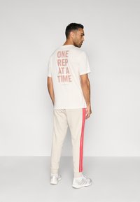 Jasnobeżowa koszulka z nadrukiem "ONE REP AT A TIME" w czerwonym kolorze na plecach, zestawiona z kremowymi dresami z czerwonymi paseczkami po bokach.