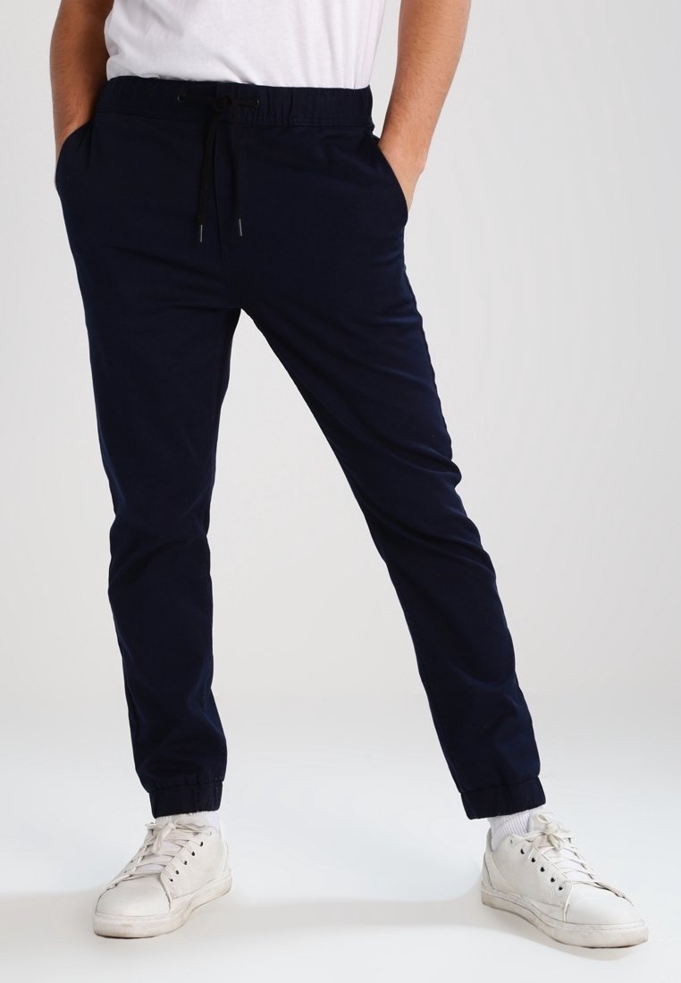 Homme portant un pantalon de jogging bleu marine avec des poignets élastiques et des baskets blanches, debout les mains dans les poches devant un fond uni.