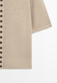 Beige gebreid kort mouw vestje met zwarte knopen langs de voorkant en een geribbelde zoom aan de mouw en onderkant.