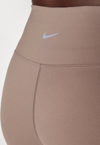 Primo piano di leggings sportivi beige a vita alta con cuciture visibili e un piccolo logo riflettente Nike swoosh vicino all'elastico in vita.