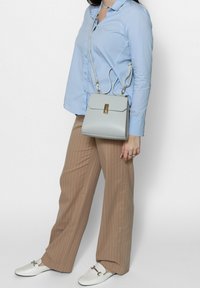 Borsa a tracolla in pelle grigia chiara con chiusura a patta e hardware dorato; abbinata a una camicia blu e pantaloni a righe beige.