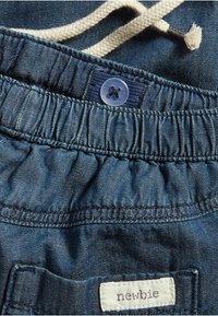 Denimshorts har ett elastiskt midjeband med dragsnöre, en ljusblå knapp och en etikett med texten "newbie" fäst vid den inre sömmen.