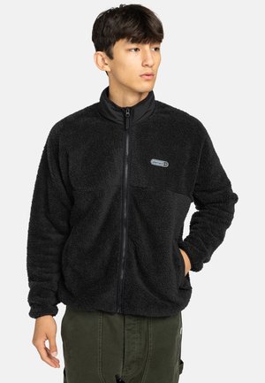Veste en polaire noire avec col haut, fermeture éclair et logo en patch sur la poitrine. Présente une texture douce et des poches avant.