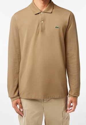 Homme portant un polo beige à manches longues avec deux boutons et un logo crocodile vert, associé à un pantalon cargo beige.