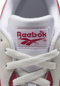 Tenisky Reebok v světle šedé semišové a bílé textilní úpravě s růžovými akcenty. Obsahují označení značky a bílé tkaničky, které se kříží přes jazyk.