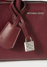 Burgundijas ādas rokassoma ar teksturētu virsmu, ar sudraba MK logo šarmu un siksnu, kurai ir birka ar iegravētu "Michael Kors".