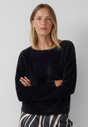 Vrouw met schouderlang blond haar, gekleed in gouden oorringen, een zwarte fuzzy trui en een zwart-beige gemusterde rok, met gekruiste armen.