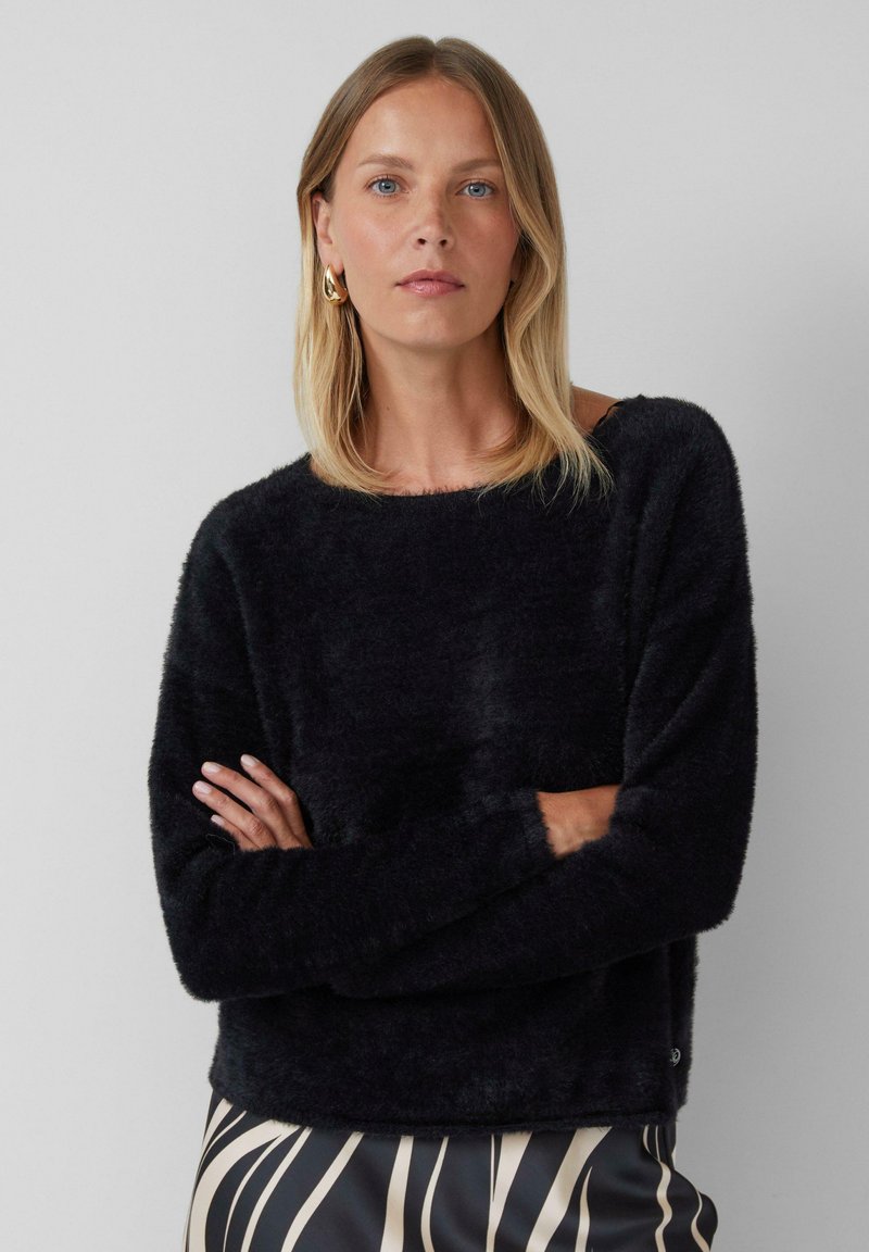 Vrouw met schouderlang blond haar, gekleed in gouden oorringen, een zwarte fuzzy trui en een zwart-beige gemusterde rok, met gekruiste armen.