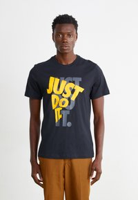 T-shirt de algodão preto com texto em amarelo e cinza em destaque "JUST DO IT". Manga curta, decote redondo e um corte descontraído. Calças castanhas usadas abaixo.