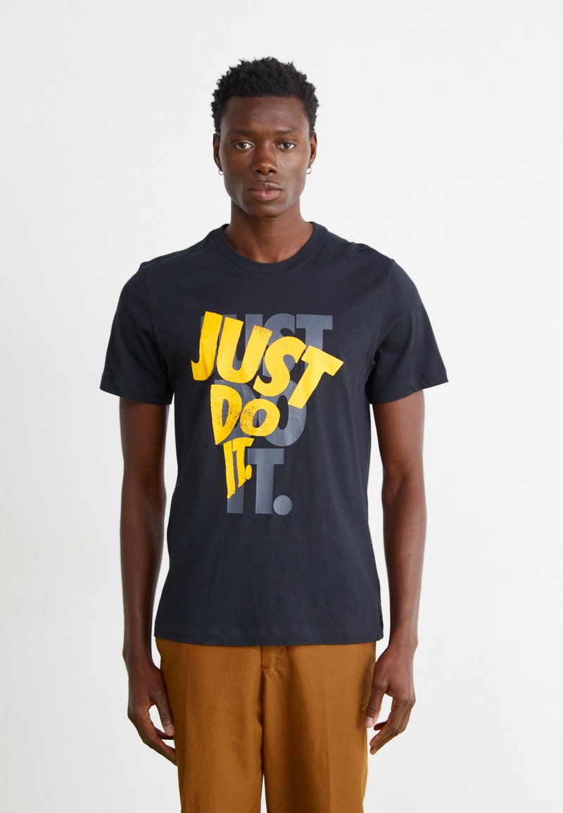 T-shirt de algodão preto com texto em amarelo e cinza em destaque "JUST DO IT". Manga curta, decote redondo e um corte descontraído. Calças castanhas usadas abaixo.