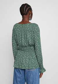 Blusa de tela texturizada con estampado floral verde, con un diseño gathers en la parte trasera y una cinturilla elástica. Presenta mangas largas con puños acampanados.