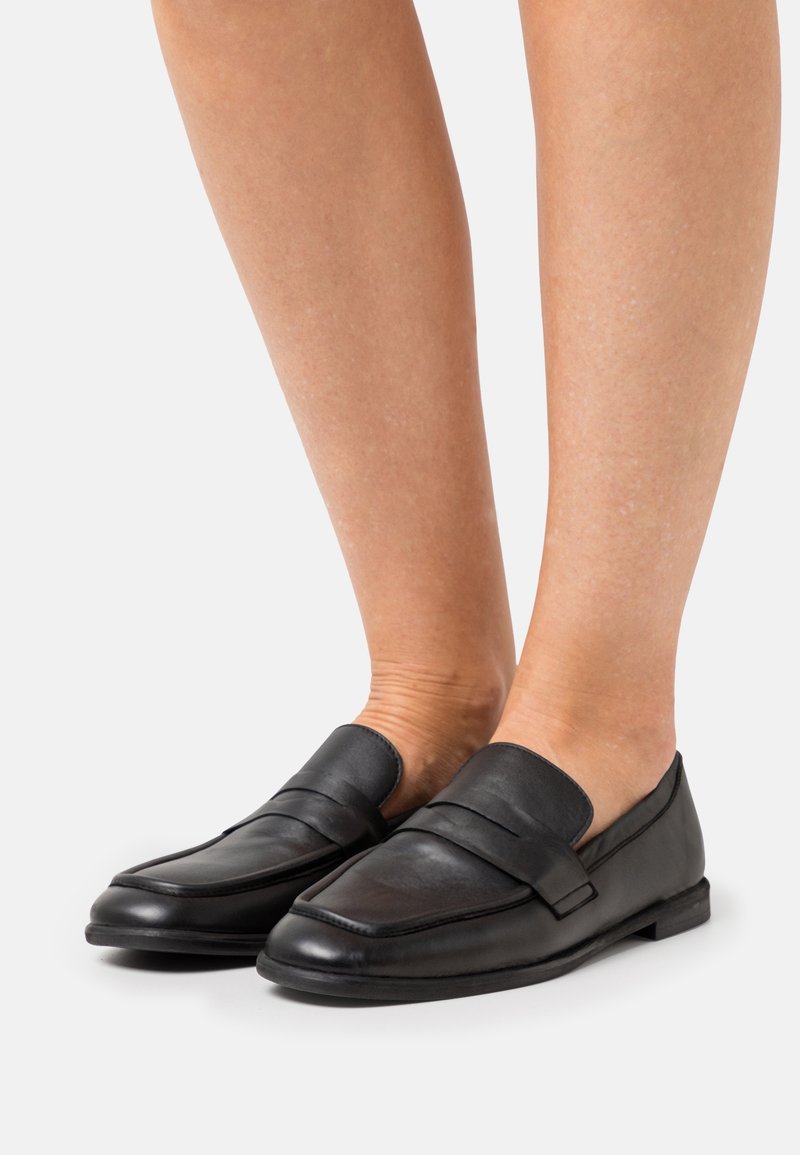 Mocassins en cuir noir à finition lisse, avec une large bride plate traversant le coup de pied et une semelle profilée basse. Pas de motifs apparents.