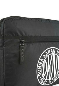 Bolsa de nylon negra con un destacado logo circular blanco. Presenta una cremallera negra y un tejido texturizado, diseñada con una forma rectangular.
