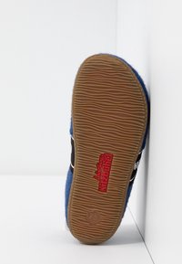 Modrá filcová bačkora s strukturovanou hnědou gumovou podrážkou, se černým popruhem a červeným brandingem "living Kitzbühel" níže. Uvedená velikost 25.