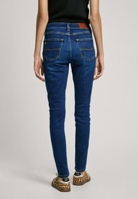 Úzke modré džíny s vysokým pásom, so zadnými vreckami s krivkovým prešívaním a hnedou koženou nášivkou na páse.