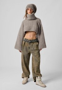 Pulover scurt gri în stil croșetat, cu guler înalt și mâneci largi; asortat cu pantaloni bej largi, cu model, și adidași albi, accentuat de o pălărie asortată.