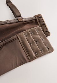 Prim-plan al unei curele de sutien maro cu trei rânduri de închizători cu cârlig și ochi și bretea de umăr reglabilă pe un fundal deschis.