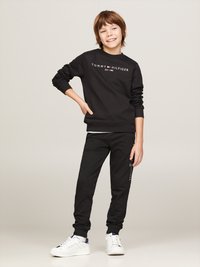 Schwarzes Sweatshirt und Jogginghosen, beide mit Logo-Akzenten. Das Sweatshirt hat einen runden Halsausschnitt und gerippte Bündchen. Weiße Sneakers runden das Outfit ab.