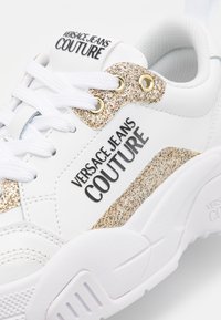 Bílé tenisky se zlatými třpytivými detaily, černým logem, texturovanou gumovou podešví, plastovými očky a zesíleným prošitím.