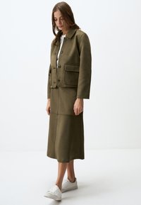Olivgrüne Jacke mit zwei vorderen Taschen, Knopfverschluss und passendem Midirock. Die Schuhe sind weiß mit dezenten grauen Akzenten.