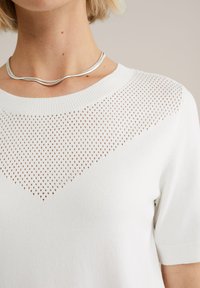 Bílý krátký svetr s krátkým rukávem a texturovaným pleteným vzorem ve tvaru diamantu na výstřihu, ribbovaným lemem a hladkým tělem. Nosit stříbrný náhrdelník.