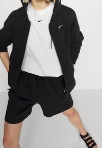 Černá mikina na zip s bílým logem Nike, bílé tričko pod ní, černé šortky a černé sandály se stahovacími pásky. Jednoduché pozadí.