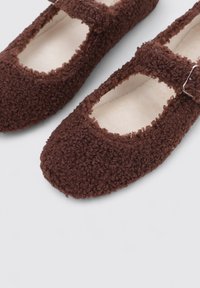 Brown fuzzy Mary Jane shoes with a textured fabric, rounded toe, and a small buckle accent at the ankle. Soft inner lining visible.