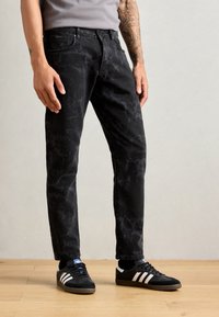 Black denim jeans with a faded, marble-like pattern, straight cut, and five-pocket design. Worn with black Adidas sneakers featuring white stripes.
