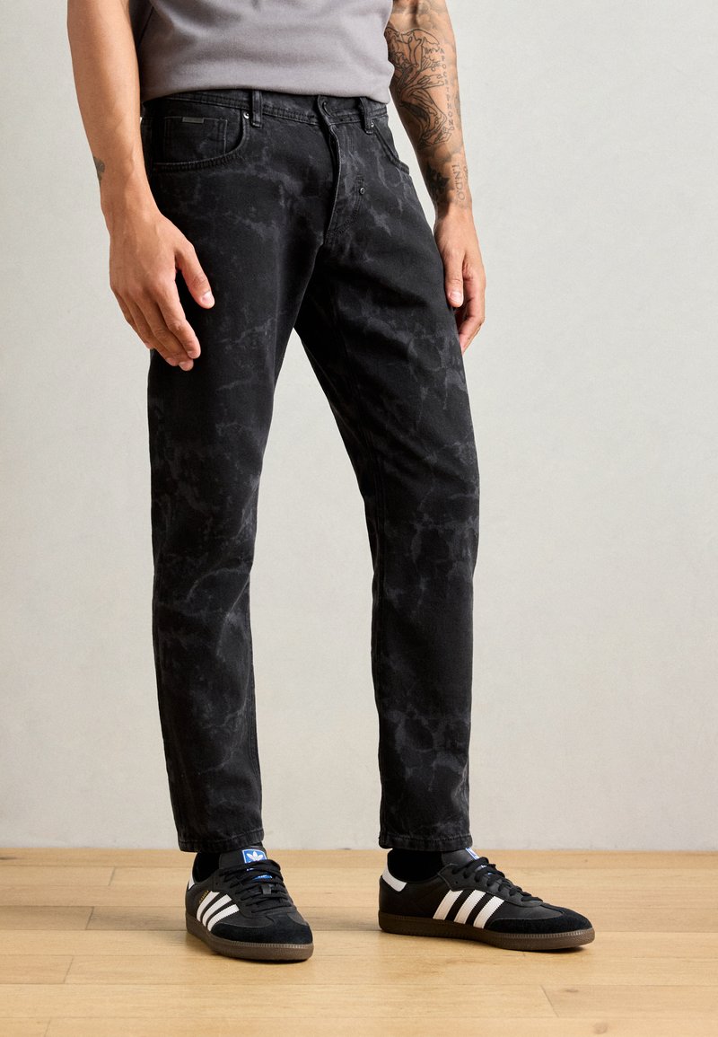 Black denim jeans with a faded, marble-like pattern, straight cut, and five-pocket design. Worn with black Adidas sneakers featuring white stripes.