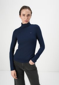 Navy geribbelde coltrui met lange mouwen, voorzien van een klein logo accent. Gecombineerd met donkergrijze broek. Vlotte textuur en slim fit.