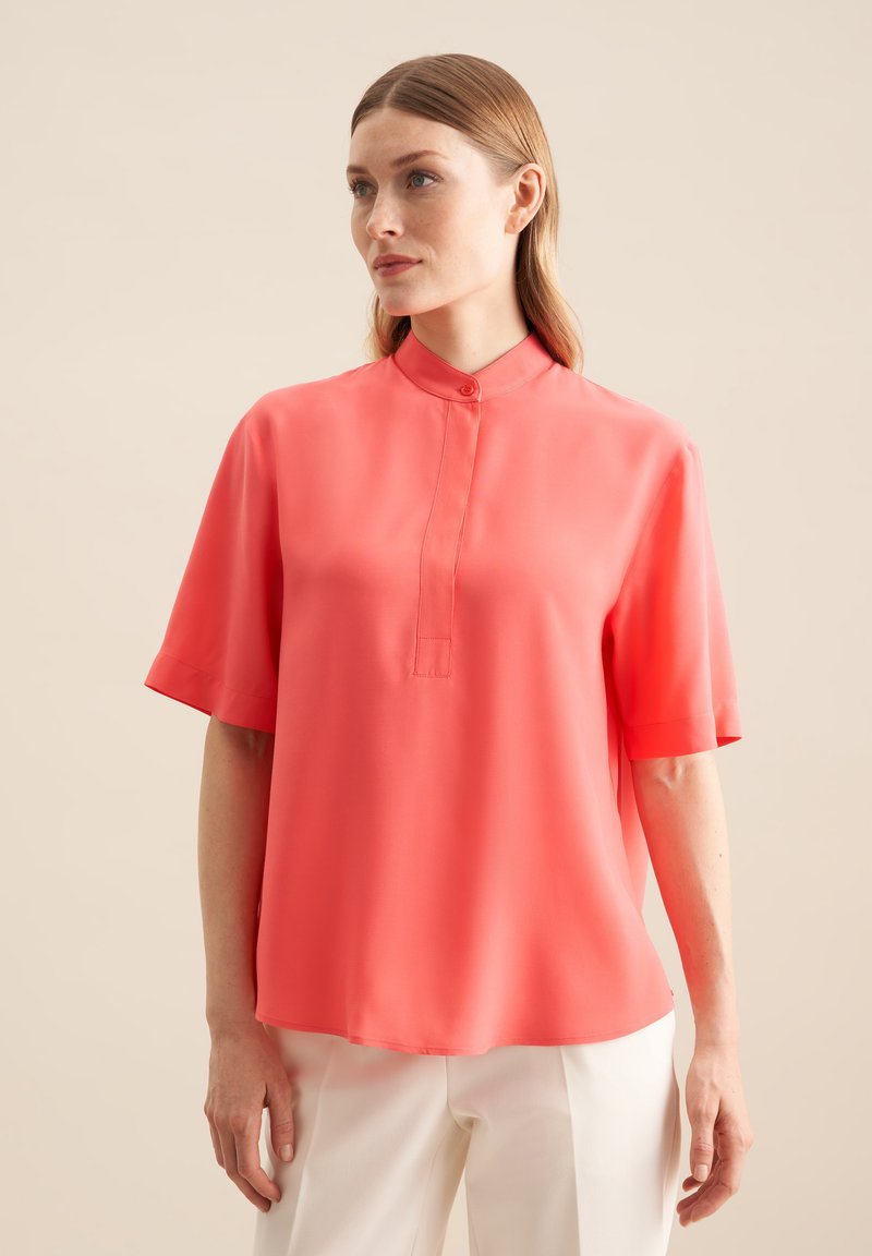 Blusa corallo a maniche corte con colletto alto, dotata di una zip frontale e realizzata in tessuto leggero e morbido. Abbinata a pantaloni bianchi.