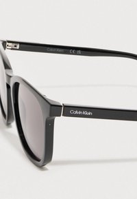 Czarne plastikowe okulary przeciwsłoneczne z ciemnymi soczewkami, kwadratową oprawą i srebrnym logo Calvin Klein na zauszniku. Widoczny metalowy zawias.