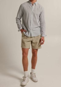 Hombre vestido con una camiseta de rayas grises y blancas de manga larga, pantalones cortos beige, calcetines blancos y zapatillas deportivas blancas, de pie contra un fondo neutral.